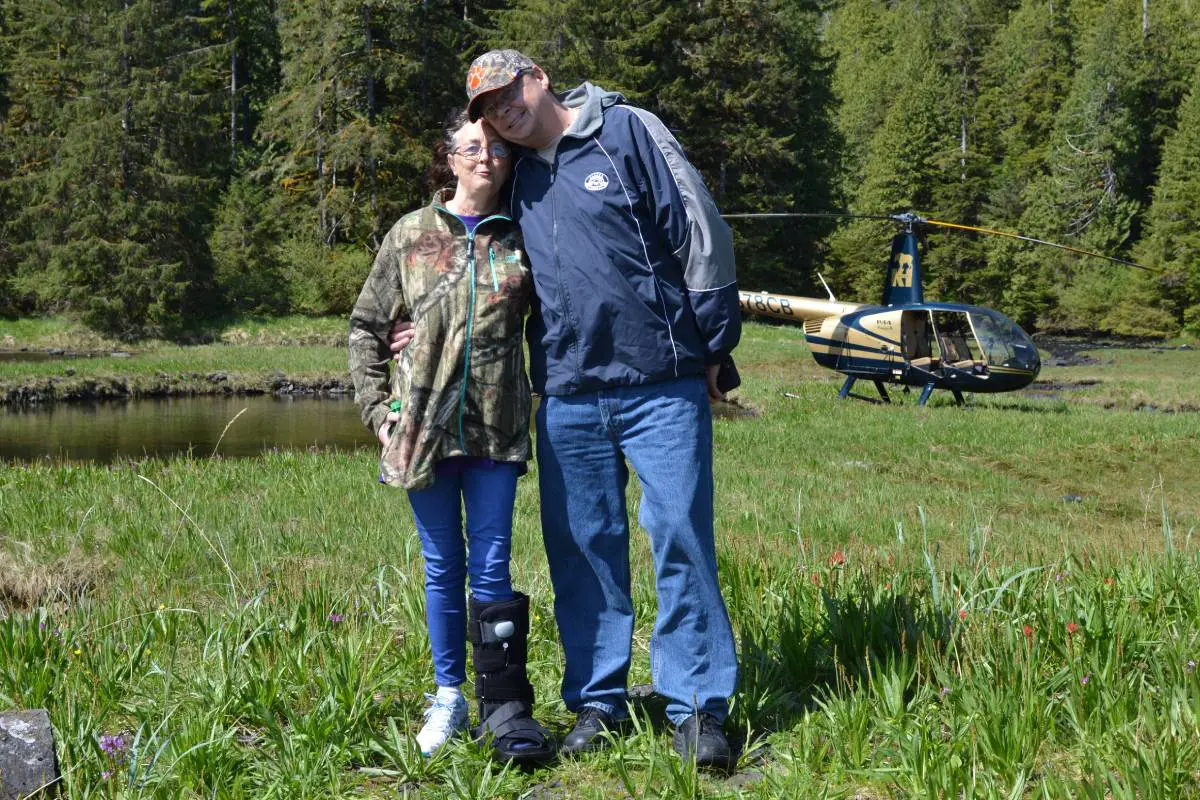 A couple stands together near a helicopter in a grassy field.